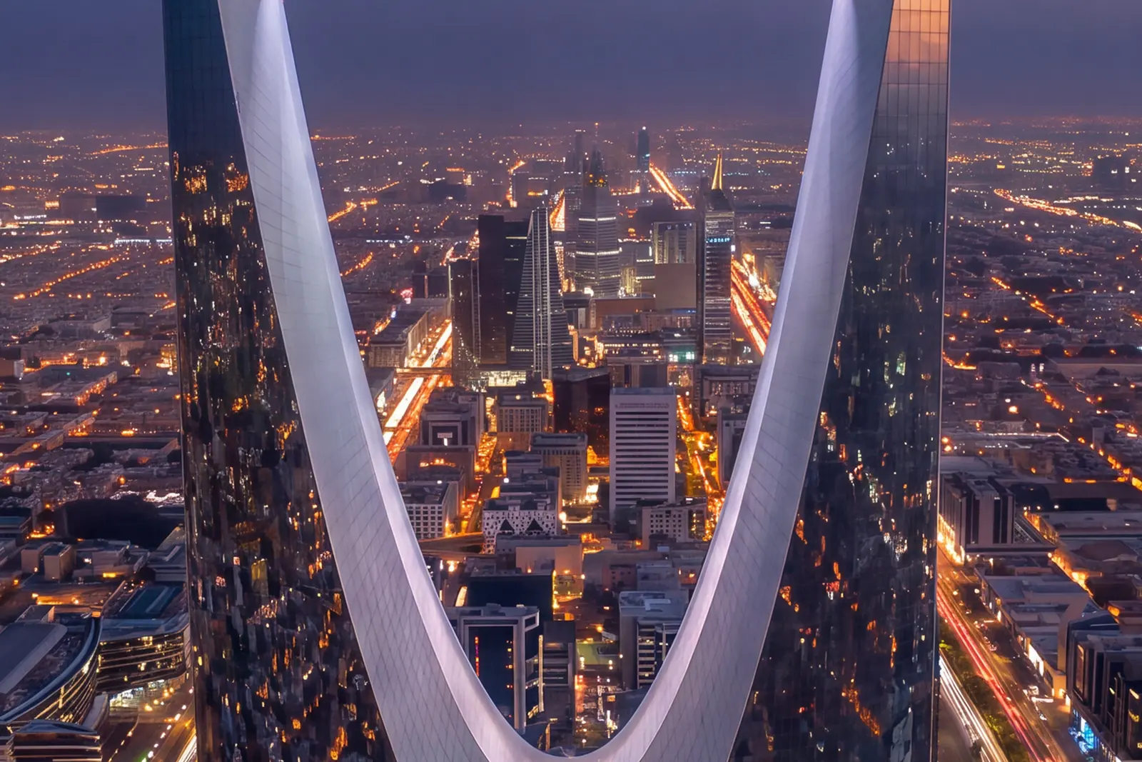 Riyadh at night from high above, framed by the arch of the Kingdom Centre: the lit grid of the city stretching to the horizon and the business district towers standing in the gap between the tower’s two faces.
