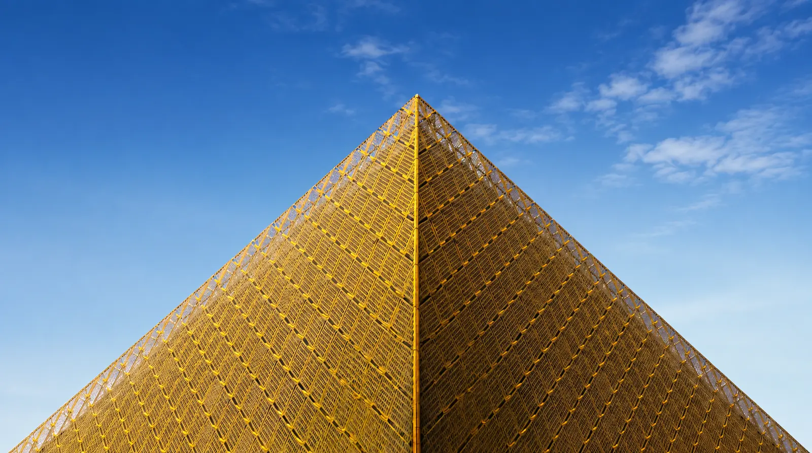 A pyramid clad in perforated brass lattice, photographed up its corner against a clear blue sky.