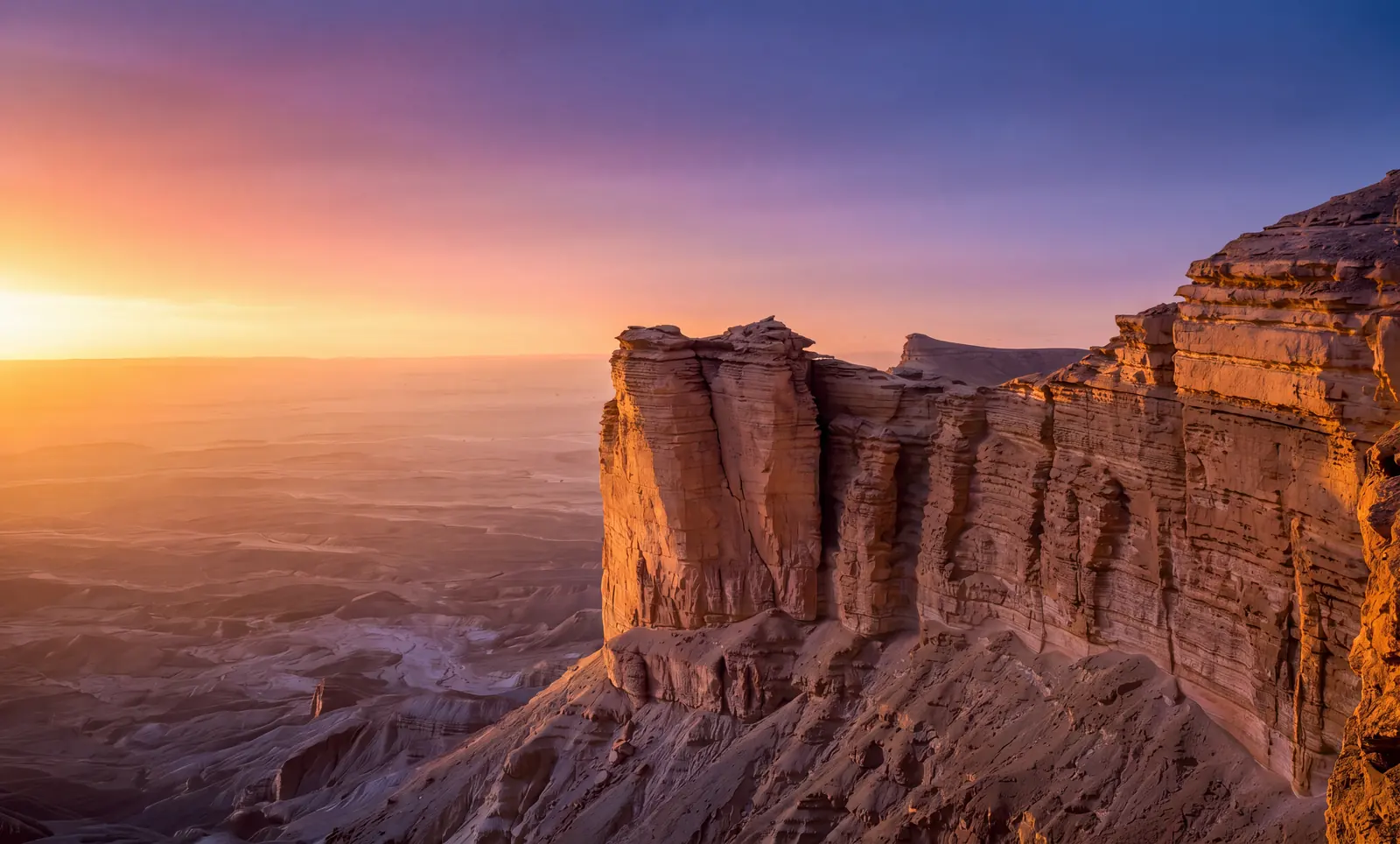 The escarpment at the Edge of the World north-west of Riyadh: sandstone pillars standing off the cliff edge above an open plain, under a sunset running from gold to violet.