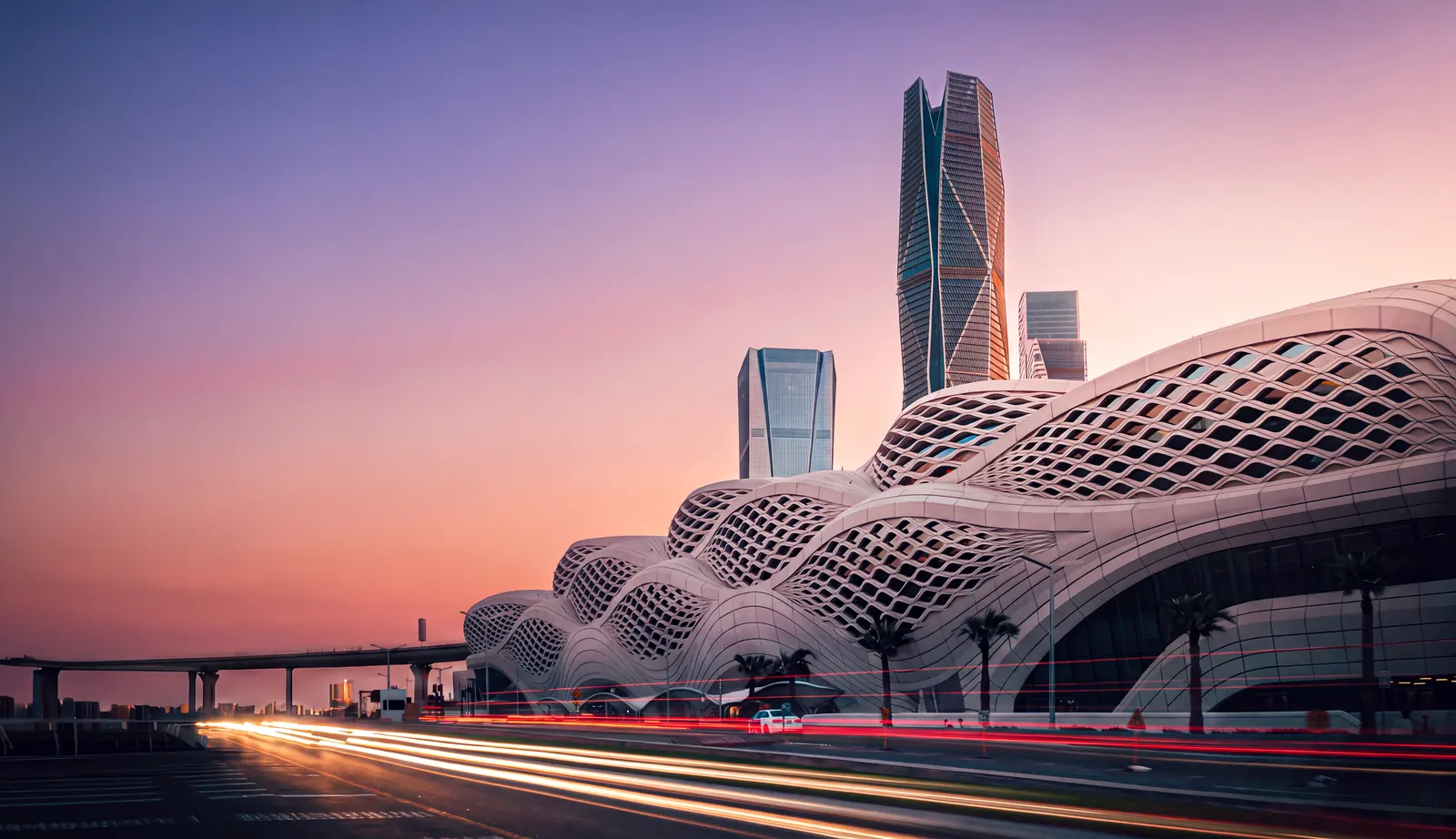 The King Abdullah Financial District metro station in Riyadh at dusk: its latticed shell under a pink sky, traffic drawn into light trails on the road in front and the district’s towers standing behind.