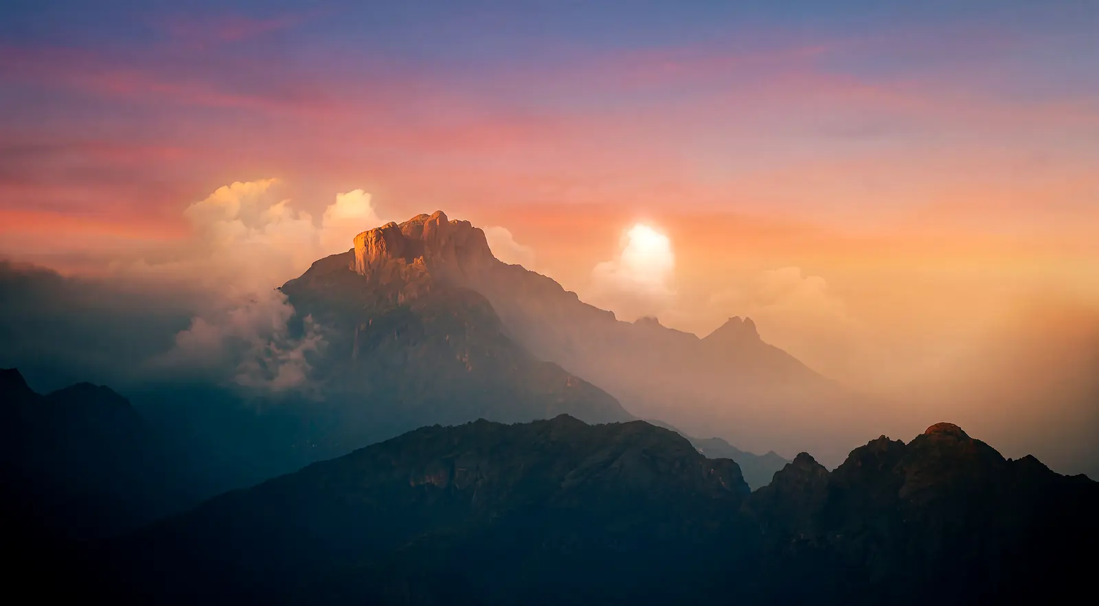 A mountain peak catching the last of the sun above a bank of cloud, the sun’s disc breaking through beside it and darker ridges falling away in front.