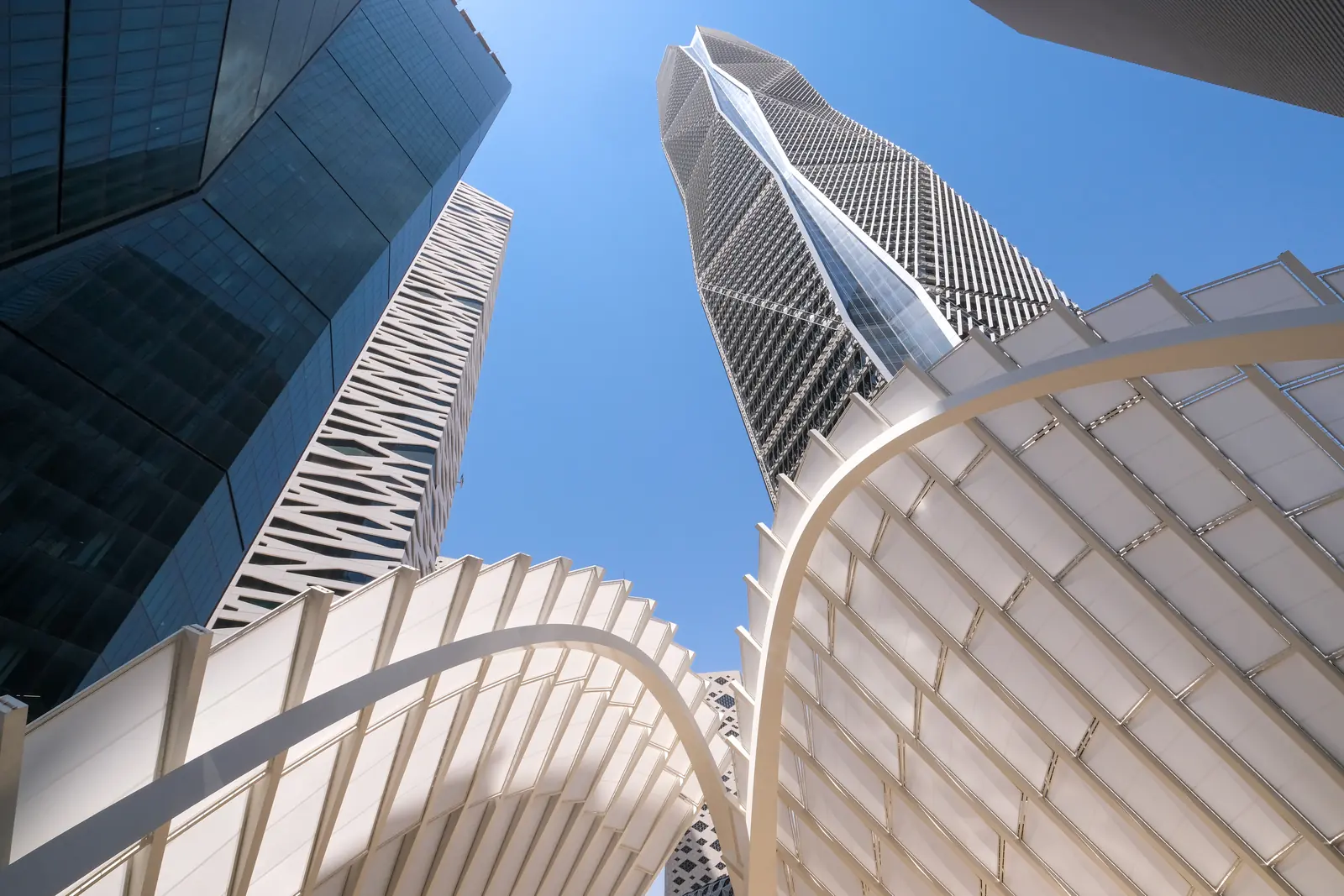 Looking straight up at the towers of the King Abdullah Financial District in Riyadh from beneath the white canopy of its plaza, the canopy’s ribs curving across a clear blue sky.