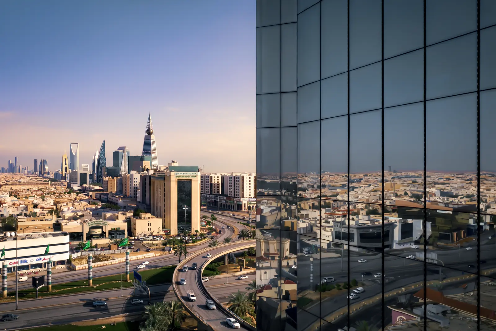 Riyadh's financial district at dusk seen past the glass flank of a tower, the city repeated across its panels.